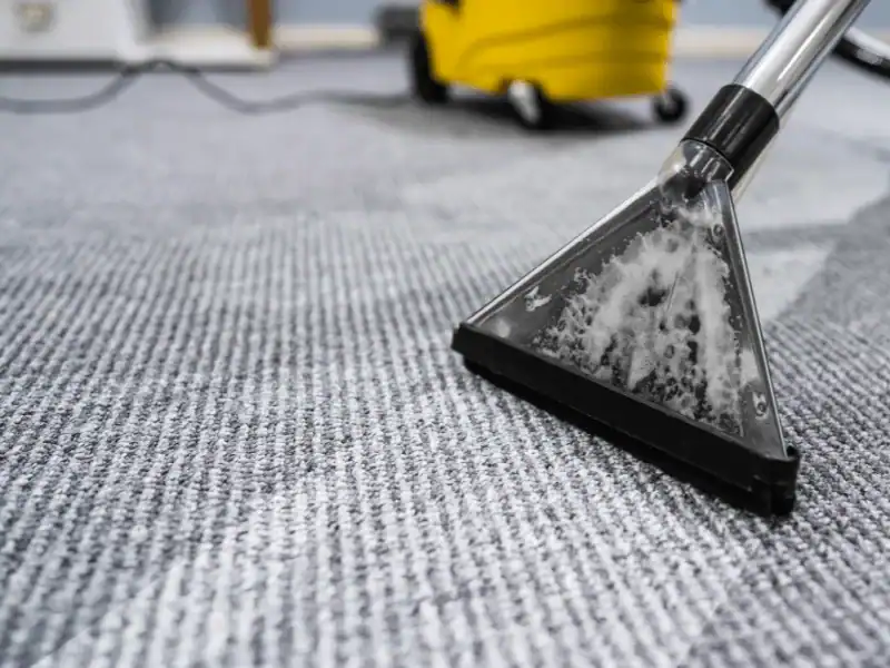 Close-up of a professional carpet extractor wand pulling steam and foam from a gray industrial carpet, showing a visible clean path being made.