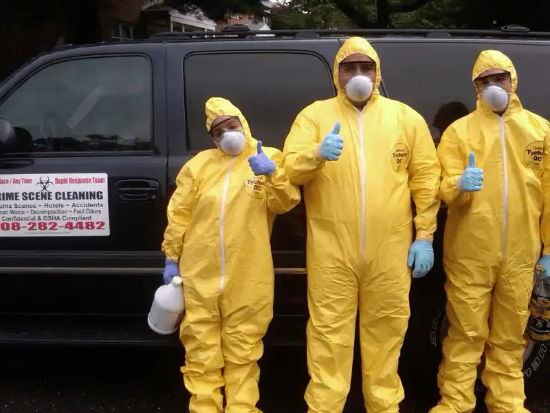 Three professionals in full yellow hazmat suits, respirators, and blue gloves giving a thumbs-up in front of a black rapid response vehicle with a biohazard warning decal.