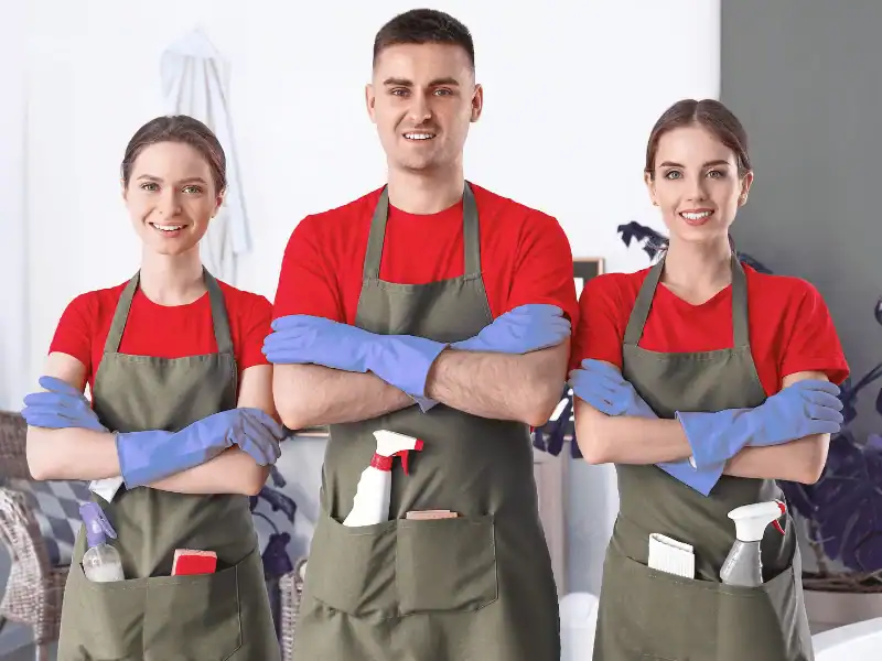 Three smiling cleaning professionals standing with arms crossed in olive green aprons and blue protective gloves, with spray bottles tucked into their apron pockets.