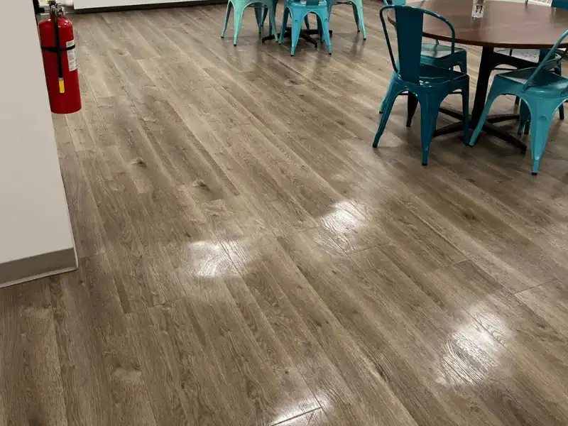 A wide shot of a high-gloss, wood-plank style vinyl floor in a commercial breakroom, showing clear light reflections on the polished surface near blue cafe chairs.