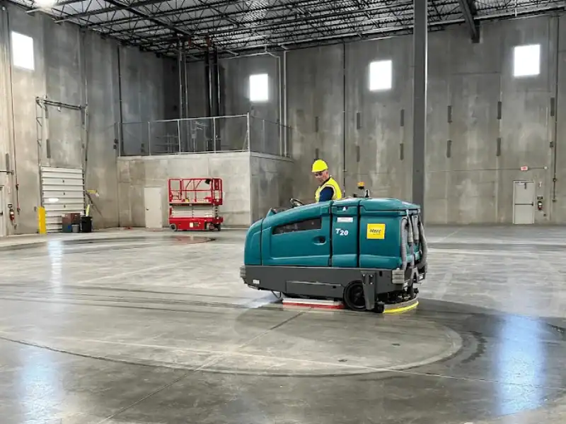 A professional operator in a yellow safety vest and hard hat driving a large teal industrial floor scrubber (T20 model) across a wide concrete warehouse floor.